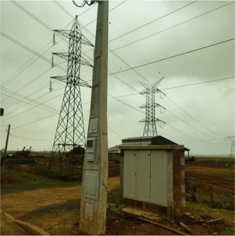 Entrada de energia em poste de concreto junto a um abrigo e torres de transmissão ao fundo